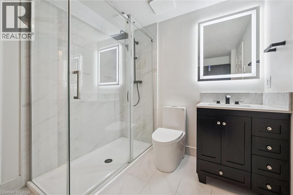 Shower with clear glass enclosure and rainfall showerhead, black vanity with an integrated sink, illuminated mirror, and a white toilet - 426 Hidden Creek Drive, Kitchener, ON