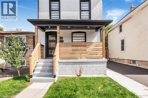 White brick facade with black trim windows, covered front porch with wood railings, modern front entry door, and concrete steps - 250 Rosslyn Avenue N, Hamilton, ON 