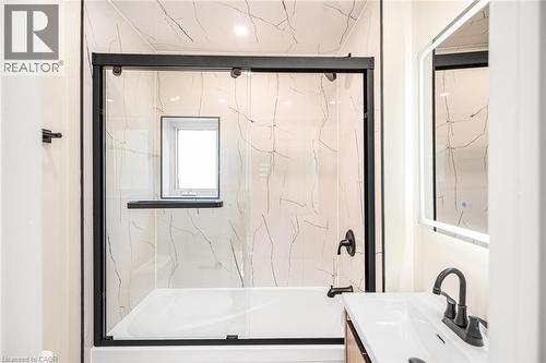 Bathtub and shower combination with black-framed sliding glass doors, white tiled walls with subtle veining, and a built-in window - 250 Rosslyn Avenue N, Hamilton, ON 