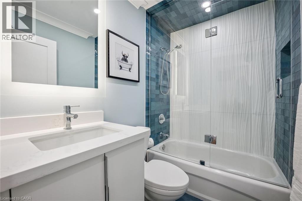 Contemporary vanity with integrated sink and chrome faucet, backlit mirror, and a shower-tub combination featuring textured white tile and blue mosaic tile accents - 1261 Stephenson Drive, Burlington, ON