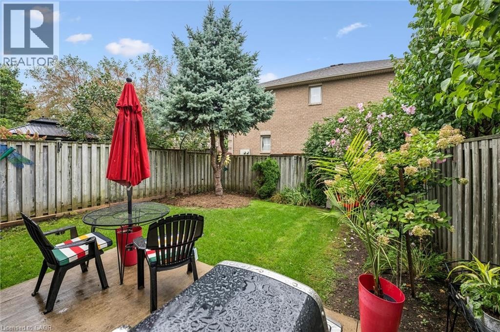 Paved patio area, manicured lawn, perimeter fencing, mature evergreen tree, and brick exterior - 1261 Stephenson Drive, Burlington, ON
