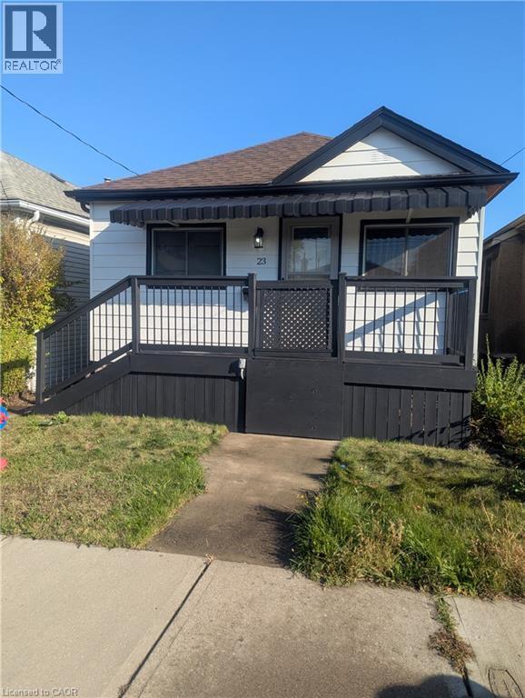 Single-story exterior featuring white horizontal siding, a gabled roofline, and a raised wooden deck with dark-finish railings - 23 Harrison Avenue, Hamilton, ON