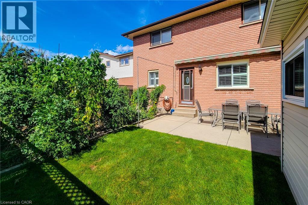 Manicured lawn, mature perimeter plantings, concrete patio, brick exterior, and a rear entry door with storm door - 276 Greencedar Drive, Hamilton, ON