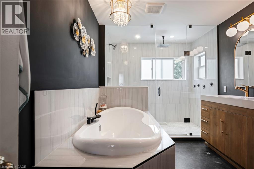 Freestanding soaking tub with a black faucet, glass-enclosed shower, wood-finish vanity with an integrated sink, and an arched mirror with backlighting - 364 Stoneheight Place, Waterloo, ON
