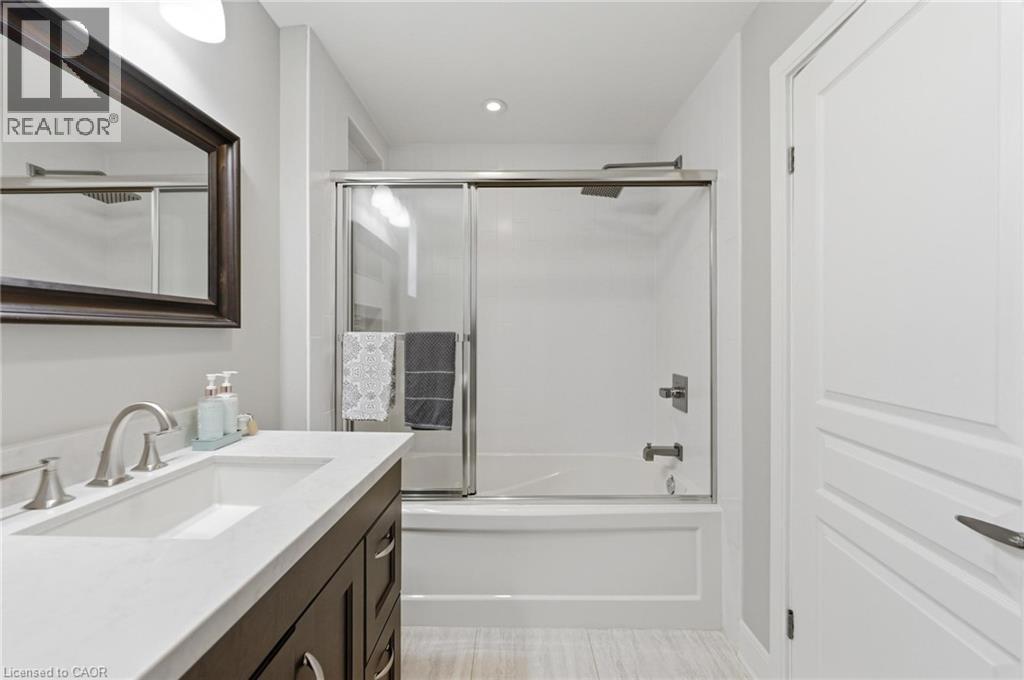 Bathroom featuring a white bathtub with a sliding glass enclosure, white rectangular sink with brushed nickel faucet, dark wood-finish vanity with drawer pulls, and a framed mirror - 69 Nash Road S, Hamilton, ON