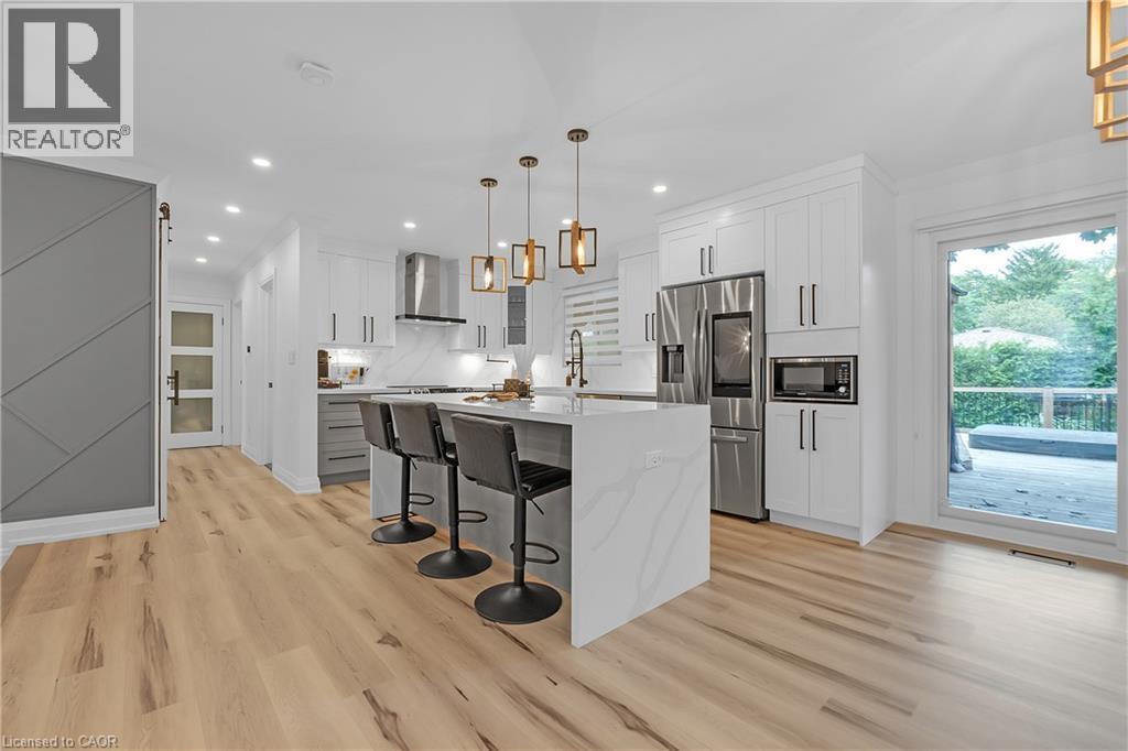 Kitchen featuring a waterfall island with integrated seating, stainless steel appliances, light-finish wood flooring, white cabinetry, and a glass sliding door - 4236 Longmoor Drive, Burlington, ON