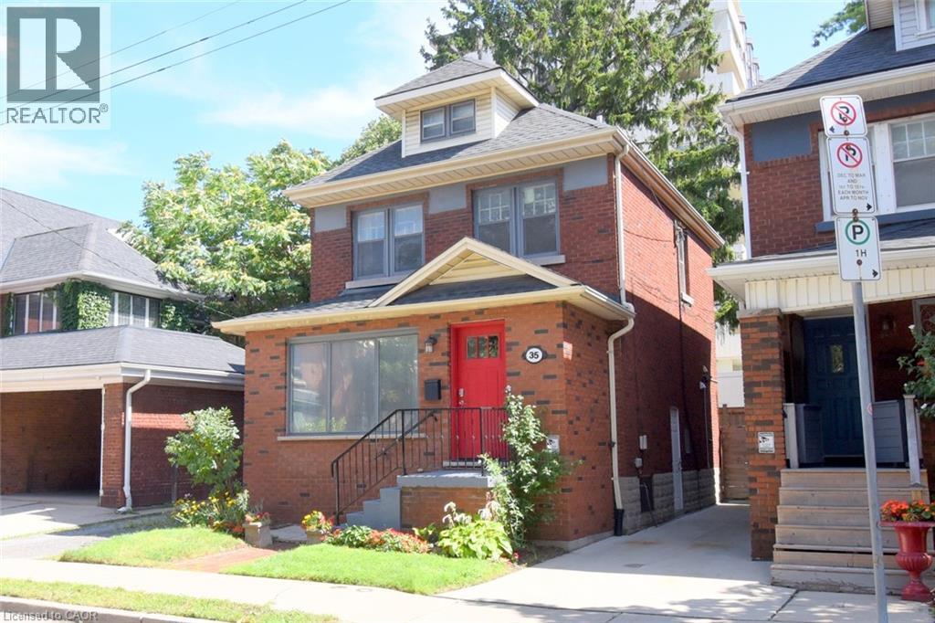 Brick exterior with a prominent red door, front porch with black railings, and a dormer window - 35 Tisdale Street S, Hamilton, ON