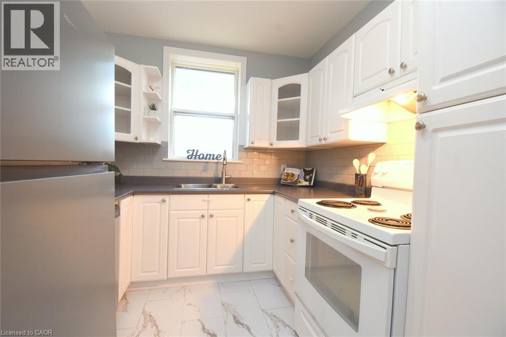 White cabinetry with brushed nickel hardware, gray subway tile backsplash, white electric range, stainless steel refrigerator, and white-patterned flooring - 35 Tisdale Street S, Hamilton, ON