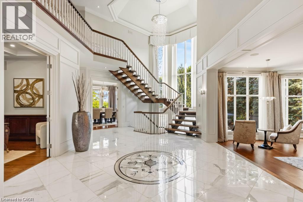Soaring ceiling with a tiered chandelier, grand staircase with wood treads and white balusters, polished tile flooring with an inlaid medallion, and expansive windows - 4038 Lakeshore Road, Burlington, ON