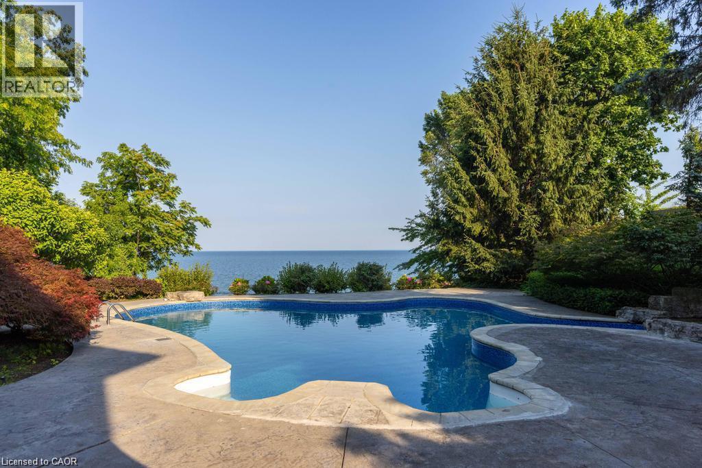 Freeform swimming pool with a stamped concrete surround, mature perimeter landscaping, and a substantial body of water in the background - 4038 Lakeshore Road, Burlington, ON