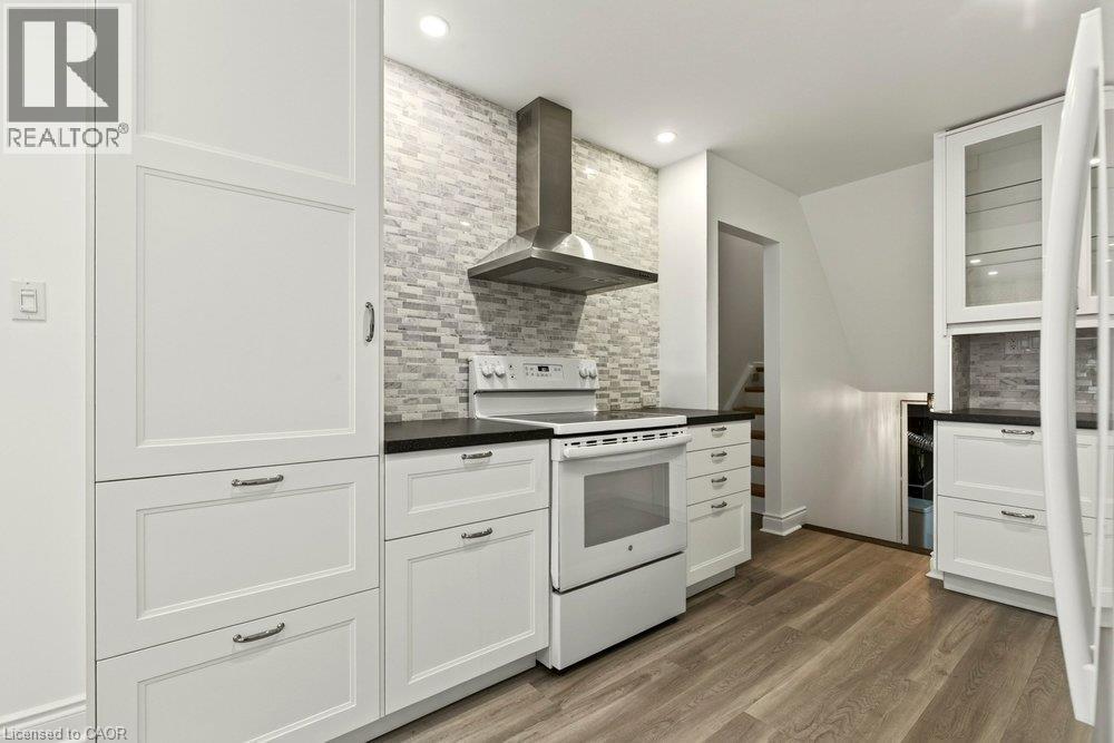 Kitchen featuring white shaker-style cabinetry, black countertops, stainless steel range hood, stone-finish backsplash, and wood-finish flooring - 27 Gretna Court, Hamilton, ON