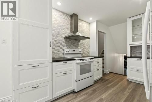 Kitchen featuring white shaker-style cabinetry, black countertops, stainless steel range hood, stone-finish backsplash, and wood-finish flooring - 27 Gretna Court, Hamilton, ON 