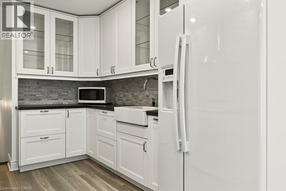 White cabinetry with glass-front uppers, dark countertops, tile backsplash, farmhouse-style sink, and wood-finish flooring - 27 Gretna Court, Hamilton, ON