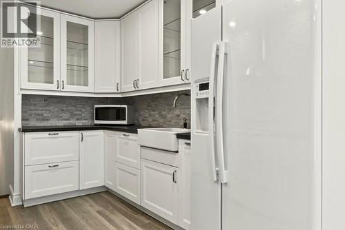 White cabinetry with glass-front uppers, dark countertops, tile backsplash, farmhouse-style sink, and wood-finish flooring - 27 Gretna Court, Hamilton, ON 