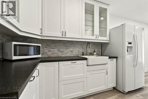 White cabinetry with glass-front upper cabinets, black countertops, a farmhouse sink, and a tile backsplash - 27 Gretna Court, Hamilton, ON 