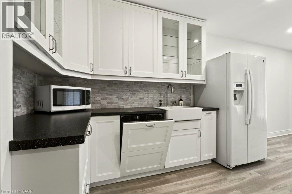 White shaker-style cabinetry with glass-front uppers, dark countertops, tile backsplash, and wood-finish flooring - 27 Gretna Court, Hamilton, ON