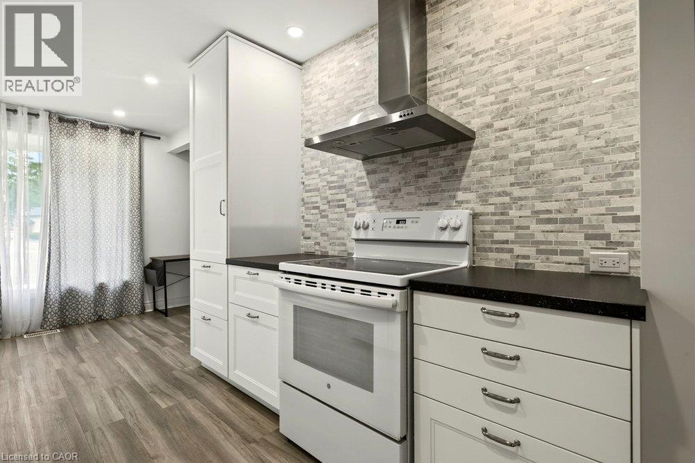 White cabinetry with dark countertops, a white electric range with a stainless steel range hood, and a light-toned brick pattern backsplash - 27 Gretna Court, Hamilton, ON