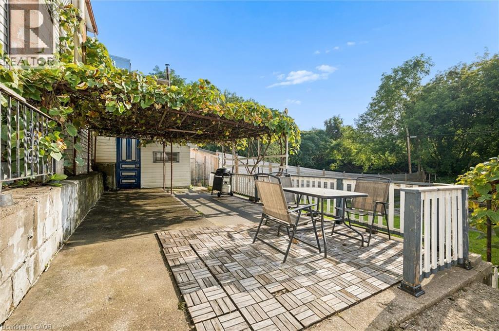 Outdoor patio area featuring a slatted wood-look floor, an overhead trellis with extensive foliage, a white picket fence, and a concrete retaining wall - 77 Beverly Street, Cambridge, ON