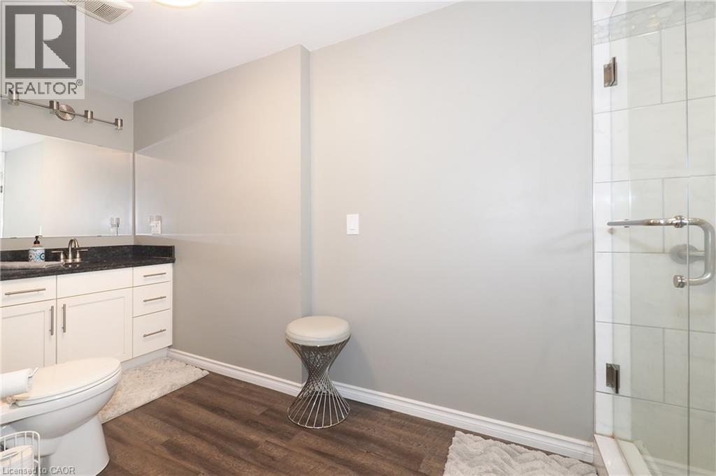Modern bathroom featuring a glass-enclosed shower, wood-finish flooring, white vanity with stone countertop, and an elongated toilet - 1200 Dunbar Road, Cambridge, ON