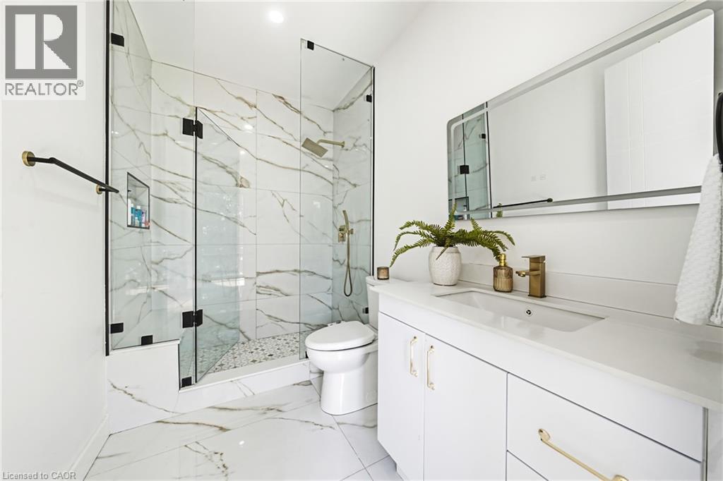Floor-to-ceiling marbled tile, glass-enclosed shower with gold-tone fixtures, white vanity with integrated sink, and a framed mirror - 4511 Guelph Line, Burlington, ON