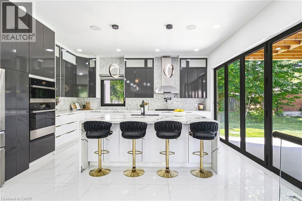 Gourmet kitchen featuring a large central island, integrated stainless steel appliances, sleek dark cabinetry, white tile flooring, and expansive folding glass doors - 4511 Guelph Line, Burlington, ON
