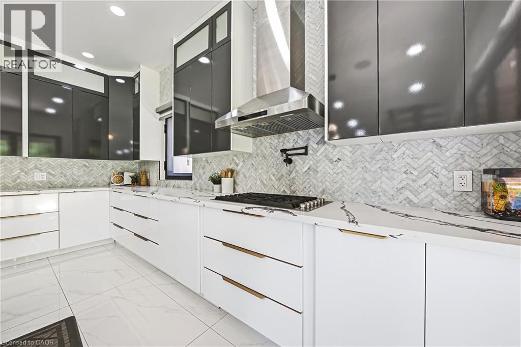 White cabinetry with gold-tone hardware, white countertops with dark veining, herringbone tile backsplash, stainless steel range hood, and dark upper cabinetry - 4511 Guelph Line, Burlington, ON