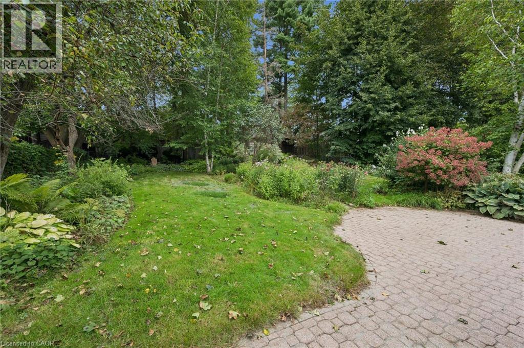 Expansive green lawn bordered by mature trees and varied plantings, with a paved brick-patterned path - 334 Coleridge Drive, Waterloo, ON