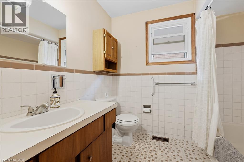 Bathroom with white tile walls, a built-in wood vanity with a white countertop and integrated sink, a toilet, and a window with wood trim - 137 Picton Street E, Hamilton, ON