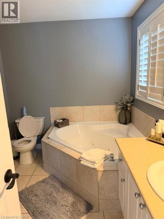 Corner jetted tub with tile surround, white plantation shutters, vanity with white cabinetry, and a toilet - 5383 Haldimand Crescent, Burlington, ON