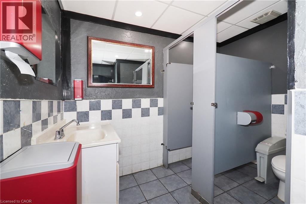 Commercial restroom featuring multiple stalls, checkerboard wall tiling, a vanity with a sink, and a framed mirror - 707 E King Street E, Cambridge, ON