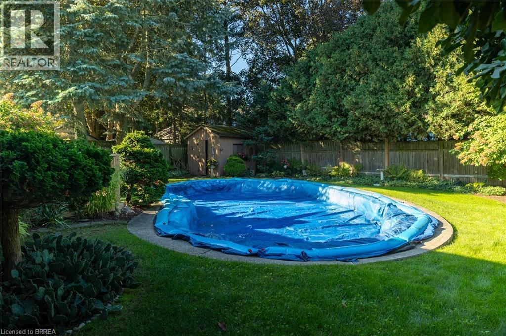 In-ground swimming pool with a concrete surround, surrounded by a manicured lawn, mature trees, and a wooden privacy fence - 78 Berkley Crescent, Simcoe, ON