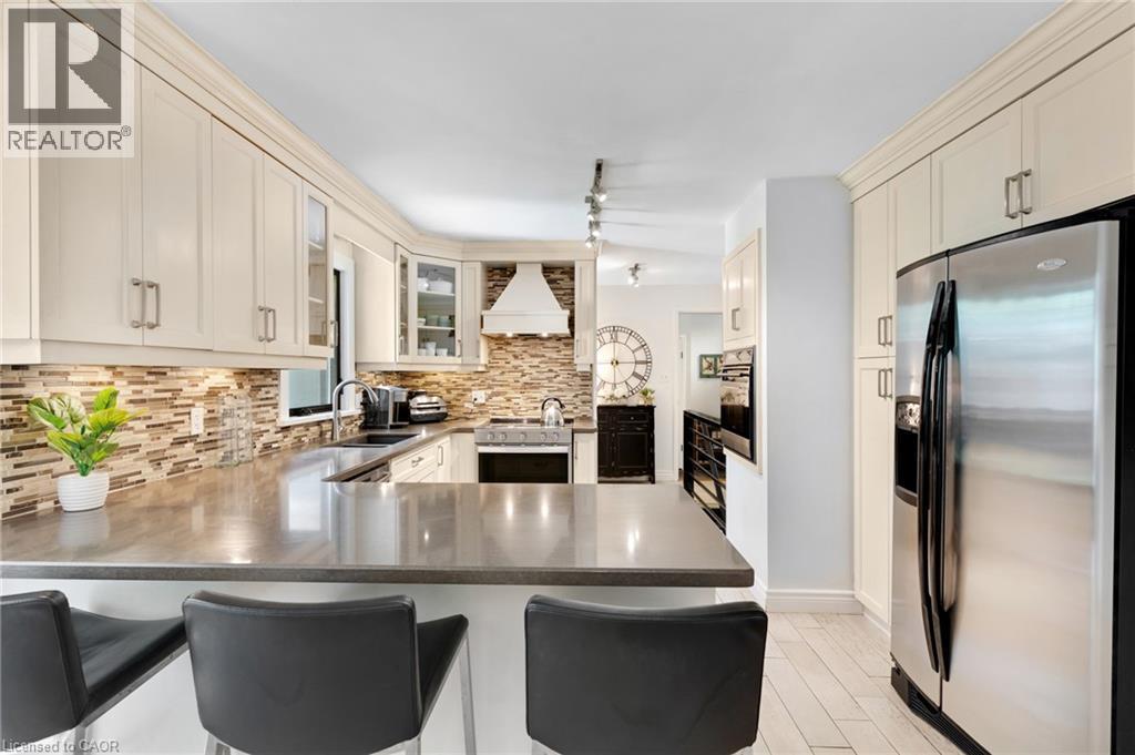 Kitchen featuring cream-colored cabinetry, stainless steel appliances, a mosaic tile backsplash, a range hood, and a built-in kitchen island with seating - 7 Shadywood Lane, Cambridge, ON