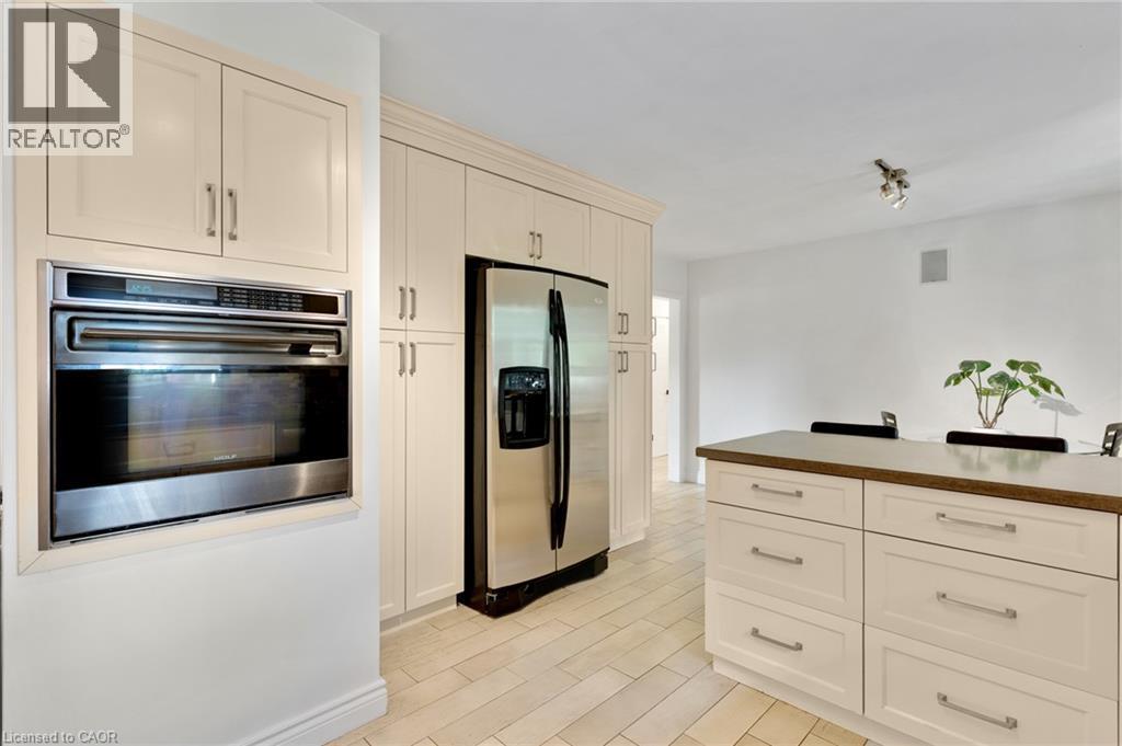 Built-in stainless steel oven, integrated refrigerator, extensive light cabinetry, kitchen island with seating, and light tile flooring - 7 Shadywood Lane, Cambridge, ON