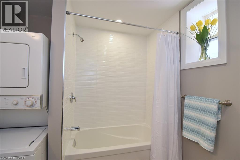 White stacked washer and dryer, white tiled shower surround, white bathtub, chrome shower fixture, and a window with white trim - 76 Burlington Street E, Hamilton, ON