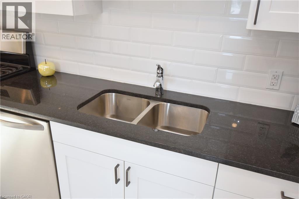 White shaker-style cabinetry with dark hardware, a dual-basin stainless steel sink, black speckled countertop, and white subway tile backsplash - 76 Burlington Street E, Hamilton, ON