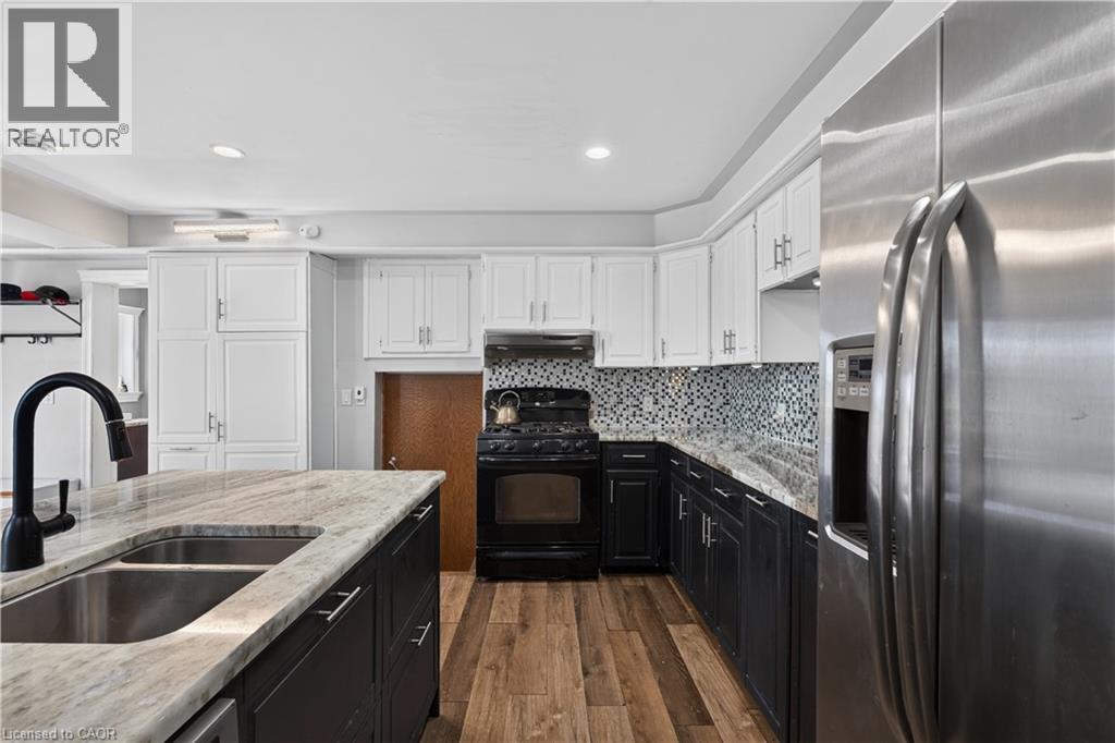 Kitchen featuring an island with a double basin sink and black faucet, wood-finish flooring, two-tone cabinetry, a tile backsplash, and stainless steel appliances - 42841 Highway 3, Wainfleet, ON