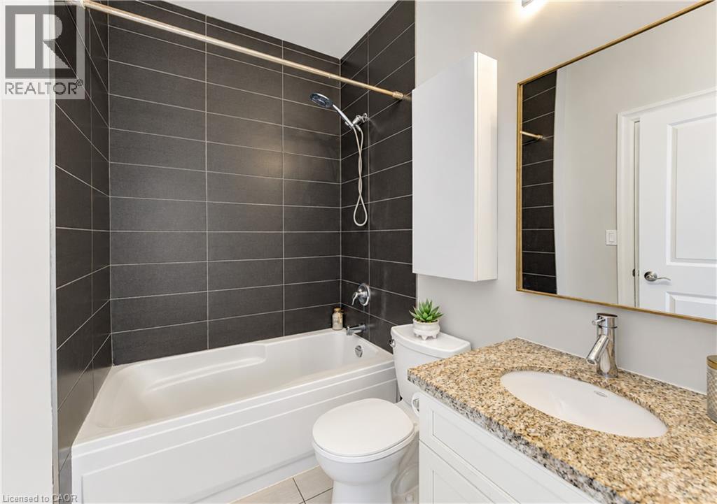 Bathroom featuring a built-in bathtub with dark tiled surround, white toilet, vanity with a light speckled countertop, and a rectangular mirror with a gold-tone frame - 25 Concession Street Unit# 110, Cambridge, ON