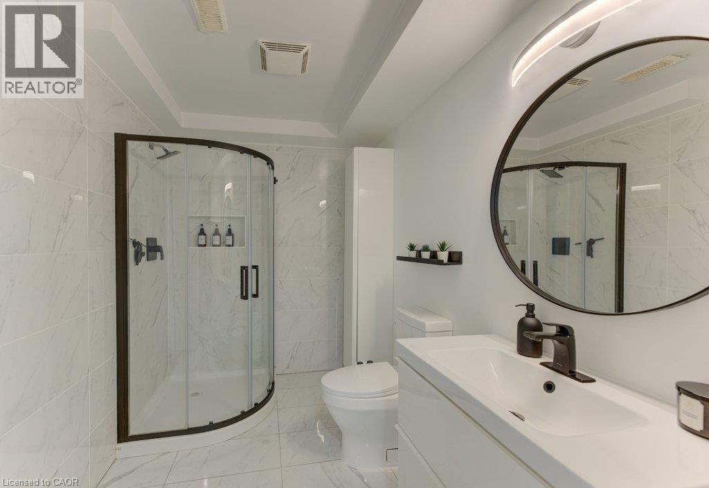 Curved glass shower enclosure with matte black framing, white tile walls and flooring, white vanity with integrated sink, matte black faucet, and a large round mirror - 88 Decaro Crescent, Cambridge, ON