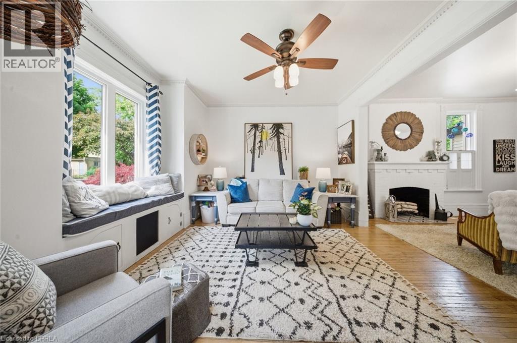 Living area featuring wood-finish flooring, a white brick fireplace, a built-in window seat with integrated storage, a ceiling fan with light fixture, and crown molding - 62 East Avenue, Brantford, ON