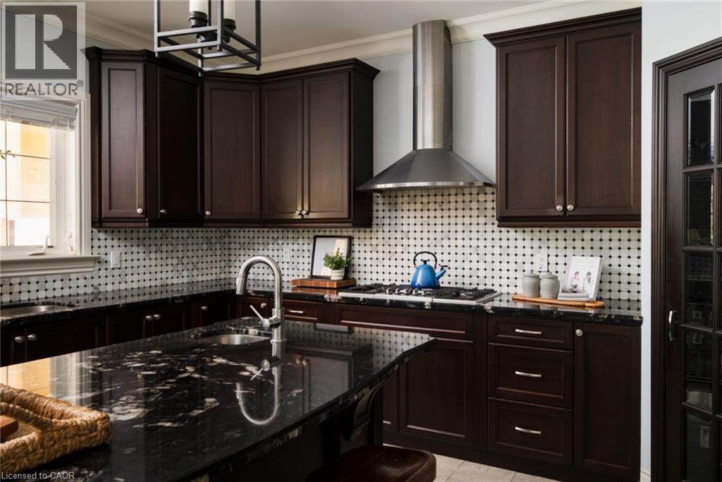 Dark wood cabinetry with crown molding, black stone countertops, a tile backsplash with black accents, a stainless steel range hood, and a built-in kitchen sink - 1505 Milburough Line, Carlisle, ON