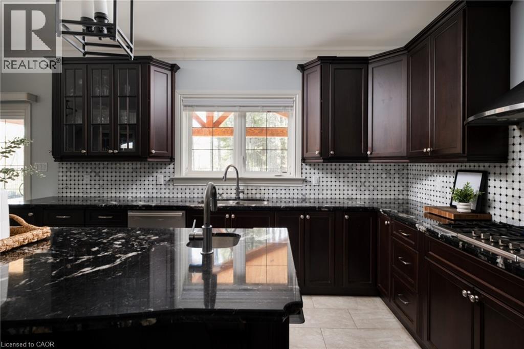 Dark wood cabinetry with glass-front uppers, stone countertops, stainless steel appliances, a tiled backsplash, and light-toned tile flooring - 1505 Milburough Line, Carlisle, ON