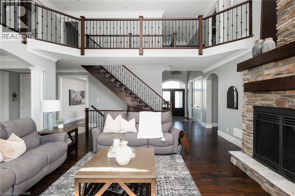 Two-story living area featuring a stone fireplace with a wood mantle, a grand staircase with wrought iron balusters, a second-story open walkway, and dark wood-finish flooring - 1505 Milburough Line, Carlisle, ON