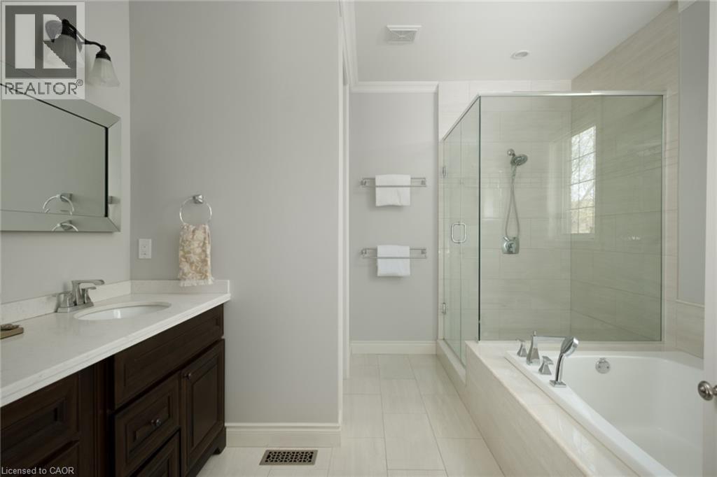 Spacious bathroom featuring a dark wood-finish vanity with a light-toned countertop, a frameless glass-enclosed shower, a built-in soaking tub, and light-toned tile flooring - 1505 Milburough Line, Carlisle, ON