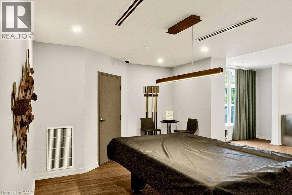Spacious recreation area featuring wood-finish flooring, a contemporary linear pendant light, recessed lighting, and a large window with floor-length drapes - 457 Plains Road Unit# 225, Burlington, ON