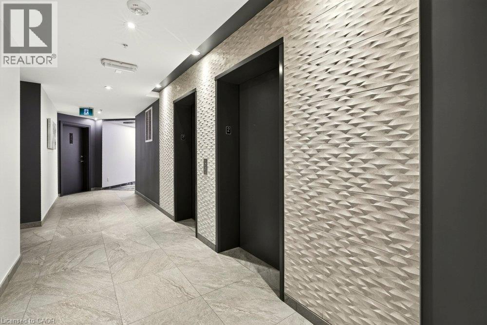 Hallway featuring two elevators with dark doors, a textured geometric accent wall, recessed lighting, and large format floor tiling - 457 Plains Road Unit# 225, Burlington, ON