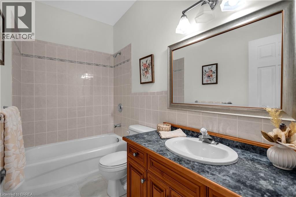 Bathroom featuring a white bathtub with light-toned tiled surround, a toilet, and a vanity with a dark speckled countertop, an oval undermount sink, and a framed mirror under a three-light fixture - 302 German School Road, Paris, ON
