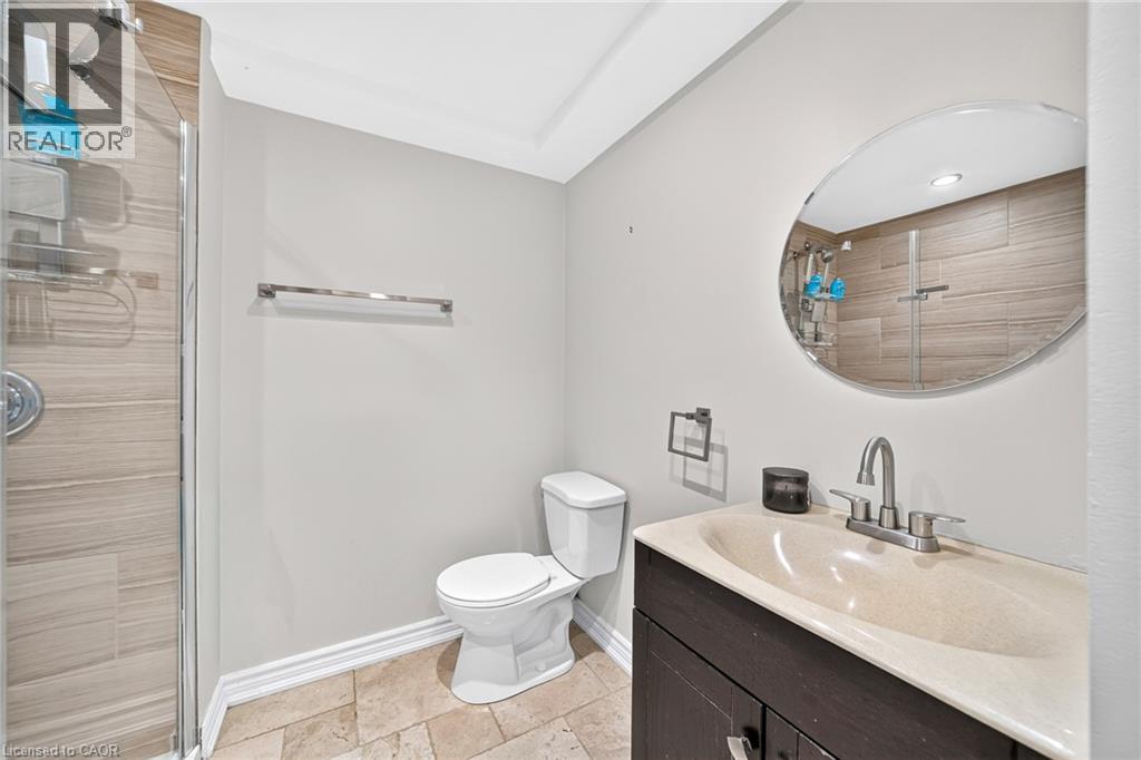 Enclosed shower with horizontal tile-finish walls, a dark wood-finish vanity with an integrated sink, a circular wall mirror, and a toilet - 1517 Elm Road Unit# 9, Oakville, ON