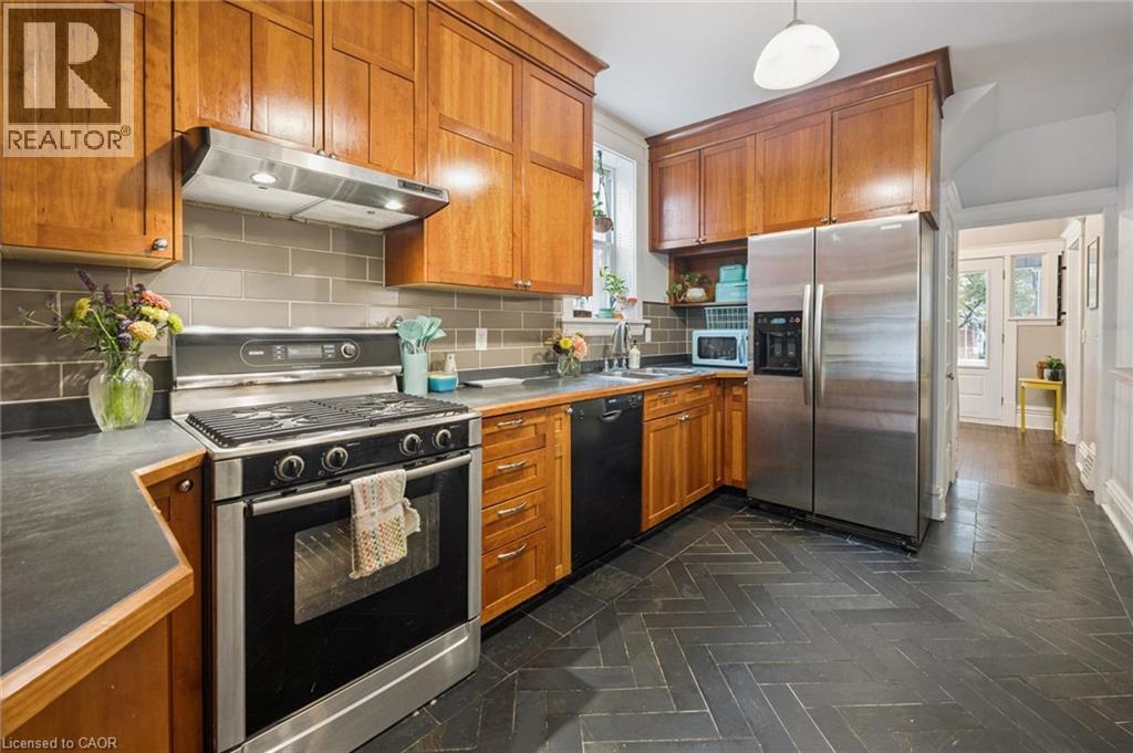Custom kitchen cabinetry with crown molding, stainless steel gas range with overhead hood fan, gray subway tile backsplash, and dark herringbone-patterned slate tile flooring - 58 Balmoral Avenue N, Hamilton, ON