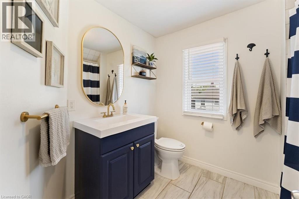 Bathroom featuring a navy blue vanity with a white countertop and gold-finish hardware, a gold-framed oval mirror, a white toilet, and light-toned tile flooring - 17 Craigmiller Avenue, Hamilton, ON