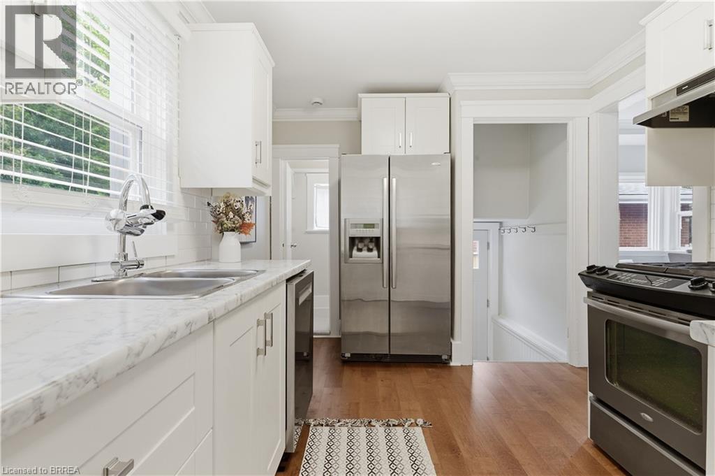 Kitchen featuring white shaker cabinetry, stainless steel appliances, wood-finish flooring, white subway tile backsplash, and a double basin sink - 87 Strathcona Avenue, Brantford, ON